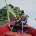 Man enjoying a peaceful boat ride near a lush island with a Jamaican flag.